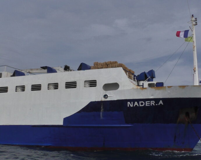 La proue d'un bateau bleu et blanc avec des bottes de foin sur le pont et l'indication "Nader-A" peinte en blanc dessus.