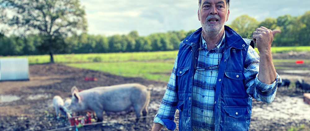 Un homme grisonnant en chemise à carreaux et gilet sans manche avec un cochon en arrière-plan.
